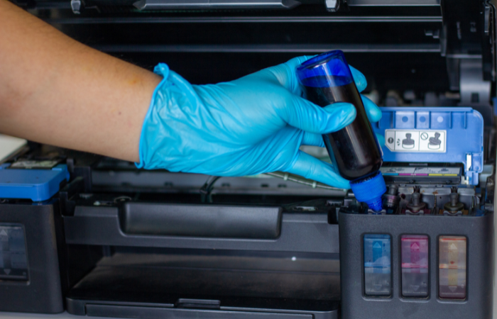  The image shows a person wearing a blue glove refilling an ink cartridge in a printer, indicating a maintenance or refill process.